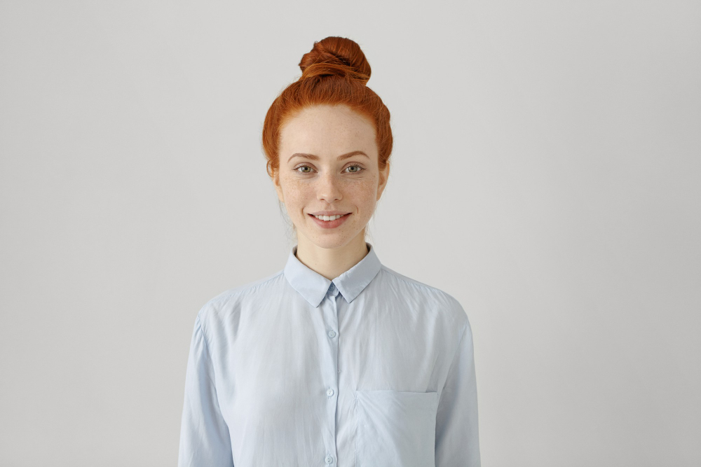 indoor shot of attractive young redhead female with hair bun in shirt smiling happily
